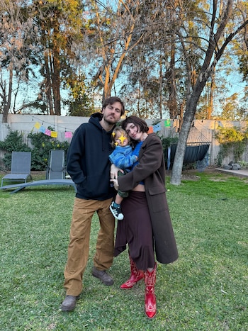 Un hombre, una mujer y un niño con el rostro cubierto por un emoji de estrella posan sobre césped, con árboles y decoraciones de papel picado al fondo
