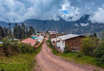 Camino de tierra ascendente en un pueblo rural andino con casas de adobe y techos de chapa. Hay árboles, arbustos verdes y montañas nubladas bajo un cielo tormentoso