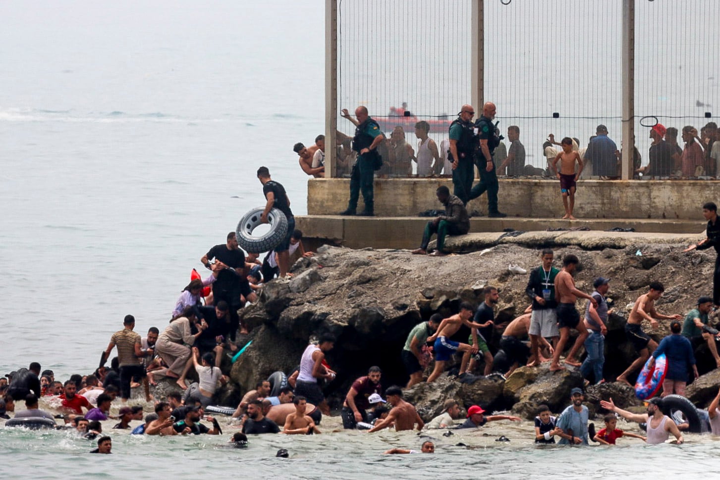 Tras alcanzar la costa, decenas de personas escalan las rocas del espigón para acceder a la ciudad. Las imágenes recuerdan a las escenas vividas durante la crisis migratoria de mayo de 2021. (EFE)
