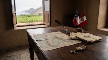 Una mesa de madera contiene un mapa antiguo, pluma, documentos, una medalla y banderas de Chile y Perú frente a una ventana abierta hacia el campo.