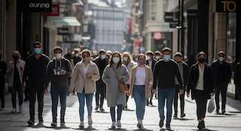 Varias personas, la mayoría con mascarillas faciales de diferentes colores, caminan por una calle urbana con edificios y tiendas a los lados.