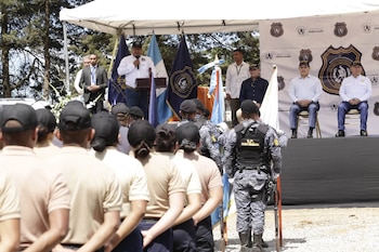 Personal uniformado de espaldas escucha a un orador en un podio bajo una carpa blanca. Banderas de Guatemala y del Sistema Penitenciario. Funcionarios sentados