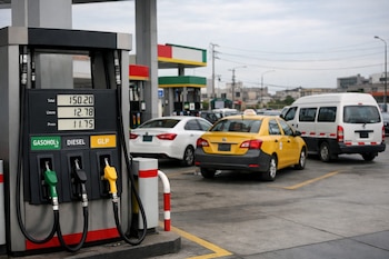 Una bomba de combustible en primer plano muestra surtidores de gasohol, diésel y GLP con precios en pantalla. Detrás, varios vehículos esperan en una estación de servicio.