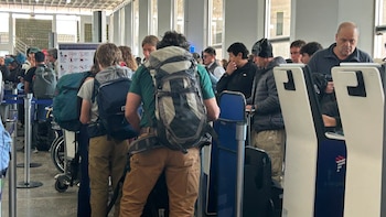 Grupo de personas con mochilas y equipaje en una fila dentro de un terminal aéreo. Se observan mostradores y carteles al fondo