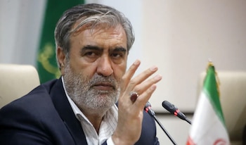 Primer plano de Ebrahim Azizi, un hombre con barba gris y traje oscuro, gesticulando con la mano derecha. Se ve un micrófono y una bandera de Irán