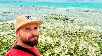 Hombre con gorra beige y barba frente a una playa de agua turquesa con algas verdes y barcos en el horizonte