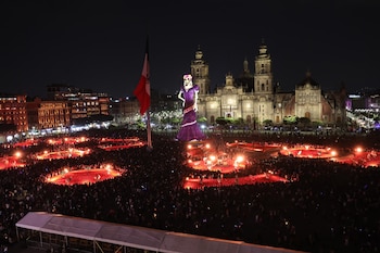 Vista nocturna de una plaza con multitud, una figura gigante con vestido morado, zonas circulares iluminadas y una catedral al fondo