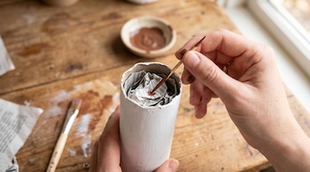 Unas manos colocan un palillo marrón dentro de un tubo de cartón relleno de periódico sobre una mesa de madera.