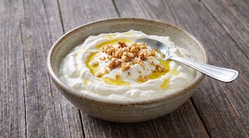 Un tazón de cerámica beige con yogur griego, nueces picadas y un líquido dorado. Una cuchara de metal plateado descansa dentro del tazón sobre una tabla de madera.