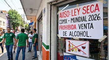 Un letrero de 'Ley Seca' en una ventana de tienda, con símbolos de bebidas alcohólicas tachadas, prohíbe su venta. Hombres con camisetas de fútbol pasan.