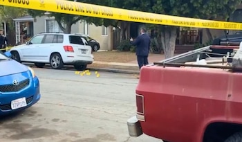 Cinta policial amarilla sobre una calle con vehículos estacionados, marcadores amarillos de evidencia en el suelo y una persona de traje