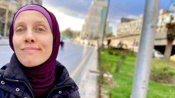 Una mujer de piel clara con un hijab morado y una chaqueta negra mira directamente a la cámara, sonriendo levemente, con una calle borrosa y edificios detrás
