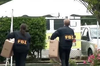 Dos personas de espaldas con chaquetas que llevan la inscripción del FBI cargan cajas de cartón frente a una carpa y vehículos