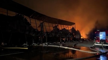 Estructura metálica en llamas durante la noche, con mangueras sobre el suelo mojado y un vehículo de bomberos a la derecha