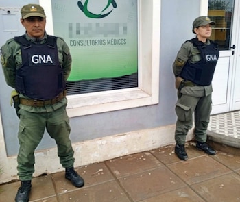 Dos agentes de Gendarmería Nacional, hombre y mujer, con uniformes verde oliva y chalecos antibalas con "GNA", de pie frente a un edificio que dice "Consultorios Médicos"
