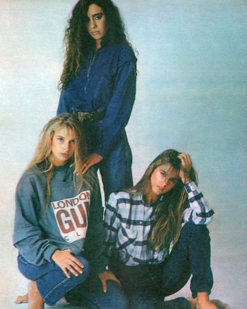 Tres mujeres posan sobre un fondo claro: una de pie con ropa azul y dos sentadas con sudadera y camisa a cuadros