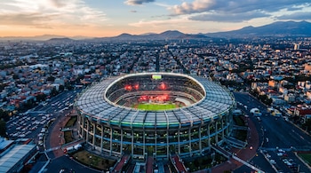 Estadio de fútbol circular iluminado con partido en juego, tribunas con público, ciudad al fondo y montañas bajo un cielo de atardecer.