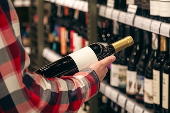 Una persona con camisa a cuadros sostiene una botella de vino con etiqueta blanca frente a estantes con diversas bebidas