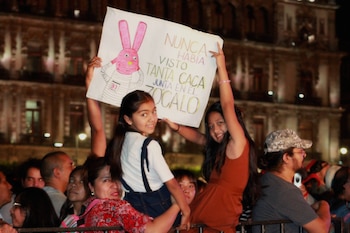 Duas garotas sorridentes segurando uma faixa branca com um coelho rosa com desenho e texto, no meio de uma multidão durante um show noturno no Zócalo