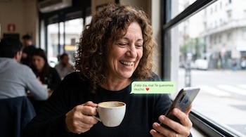Una mujer de cabello rizado sostiene una taza blanca y mira la pantalla de un teléfono móvil que muestra un mensaje junto a una ventana.