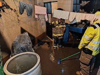 En un patio inundado de cemento, dos personas limpian barro y agua; una es bombero, otra lleva chaqueta amarilla. Ropa colgando en tendedero