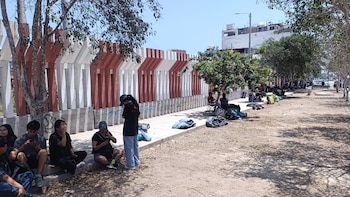 Varias personas sentadas en una vereda junto a una reja de concreto, árboles y bolsas con pertenencias