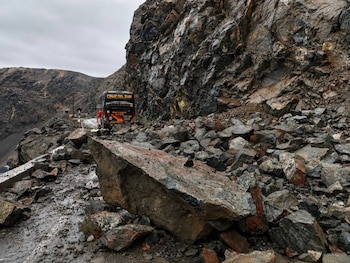 Una carretera cubierta de rocas y tierra con un autobús rojo y amarillo detenido en el fondo, junto a una ladera rocosa bajo un cielo nublado