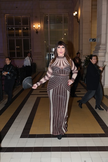 Moria Casán de pie, sonriendo, con un vestido largo ajustado de rayas marrones y beige brillantes, sosteniendo un abanico negro. Fondo de edificio con seguridad