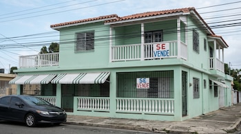 Fachada de una vivienda caribeña de dos pisos color verde menta con balcón blanco, rejas negras y toldos. Carteles "SE VENDE" visibles. Coche negro aparcado en la calle.