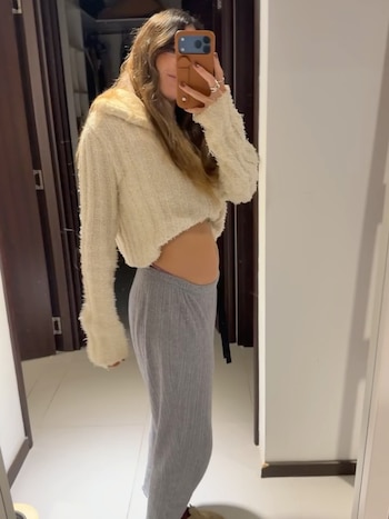 Mujer de cabello largo castaño se toma una selfie en el espejo con celular, vistiendo un suéter corto color crema y pantalón gris de punto