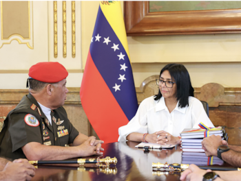 Una mujer con lentes y camisa blanca conversa sentada a una mesa con un militar con uniforme y boina roja ante una bandera de Venezuela