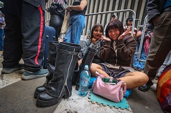 Dos jóvenes sentadas en el suelo junto a unas botas altas de color negro, una botella de agua, un bolso rosado y vallas metálicas