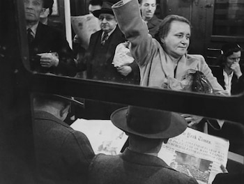 Fotografía en blanco y negro de pasajeros en un vagón de metro, incluyendo una mujer con el brazo alzado y un hombre leyendo el periódico New York Times