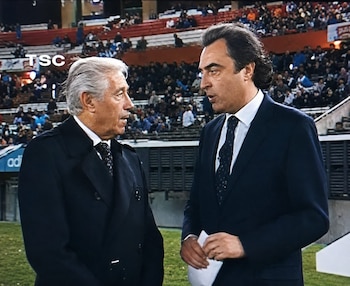 Dos hombres de traje conversan en el borde de un campo de fútbol frente a una tribuna con público en un estadio
