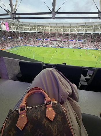 Vista desde un asiento de estadio de fútbol. El campo verde con jugadores en uniformes rosados y negros. En primer plano, piernas y un bolso de diseño sobre el regazo