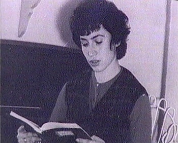 Una mujer joven con cabello rizado oscuro y corto está sentada leyendo un libro abierto, vestida con una camisa y un chaleco