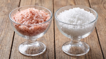 Dos cuencos de vidrio transparentes llenos de sal gruesa; uno contiene sal rosa del Himalaya y el otro sal blanca marina sobre una superficie de madera.