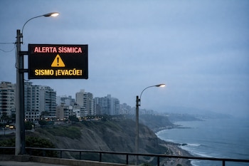 Cartel digital con texto 'ALERTA SÍSMICA' y 'SISMO ¡EVACÚE!', símbolo de advertencia, dos postes de luz, edificios, acantilados, carretera con vehículos, Océano Pacífico y cielo.