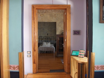 Una puerta de madera se abre a un dormitorio con cama de hierro forjado. A la derecha, una mesa de madera con libros y una silla