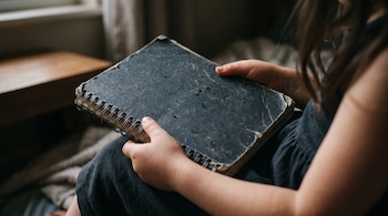 Manos de una niña que sostienen un cuaderno cerrado con espirales y tapas oscuras desgastadas.