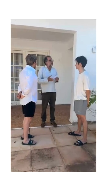 Tres hombres de pie en un patio con baldosas. Uno de espaldas con camisa blanca, otro con barba gesticulando y un joven con camiseta blanca