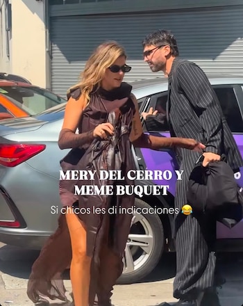 Una mujer con vestido marrón y gafas oscuras camina junto a un hombre con traje a rayas que sostiene un bolso frente a un automóvil