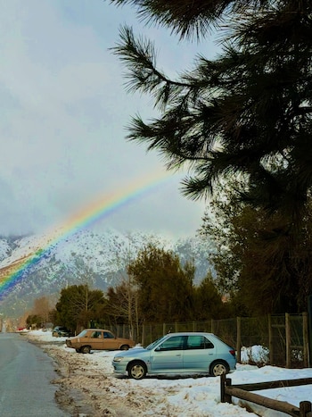 Cielo nublado con arcoíris sobre montaña nevada. Primer plano de pinos, carretera asfaltada con nieve y dos vehículos estacionados