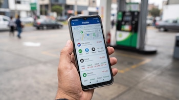 Una mano sostiene un teléfono móvil que muestra la aplicación Facilito con precios de combustible frente a un surtidor de gasolina.