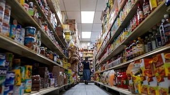 Una persona camina por el centro de un pasillo estrecho de supermercado flanqueado por estantes altos y repletos de productos alimenticios envasados