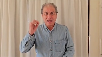 Hombre de cabello gris y camisa de mezclilla clara hablando, con la mano derecha levantada, frente a una cortina de color crema