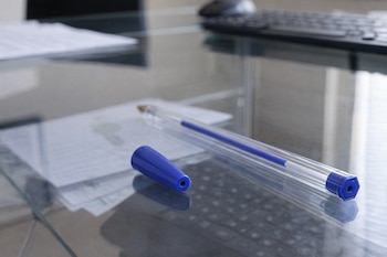 Primer plano de un bolígrafo azul de plástico transparente con su tapa azul separada y con agujero, colocado sobre una mesa de cristal con documentos.
