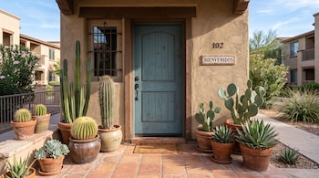Entrada de vivienda con una puerta de madera azul, el número 102, un letrero de Bienvenidos y diversas macetas con cactus alrededor.