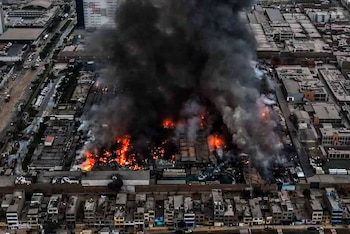 Vista aérea muestra un incendio con llamas brillantes y una densa columna de humo negro elevándose desde una planta industrial en una zona urbana con edificios y calles