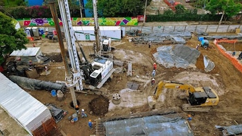 Vista aérea de un terreno en obra con una excavadora amarilla, una perforadora de suelo, contenedores y obreros con cascos